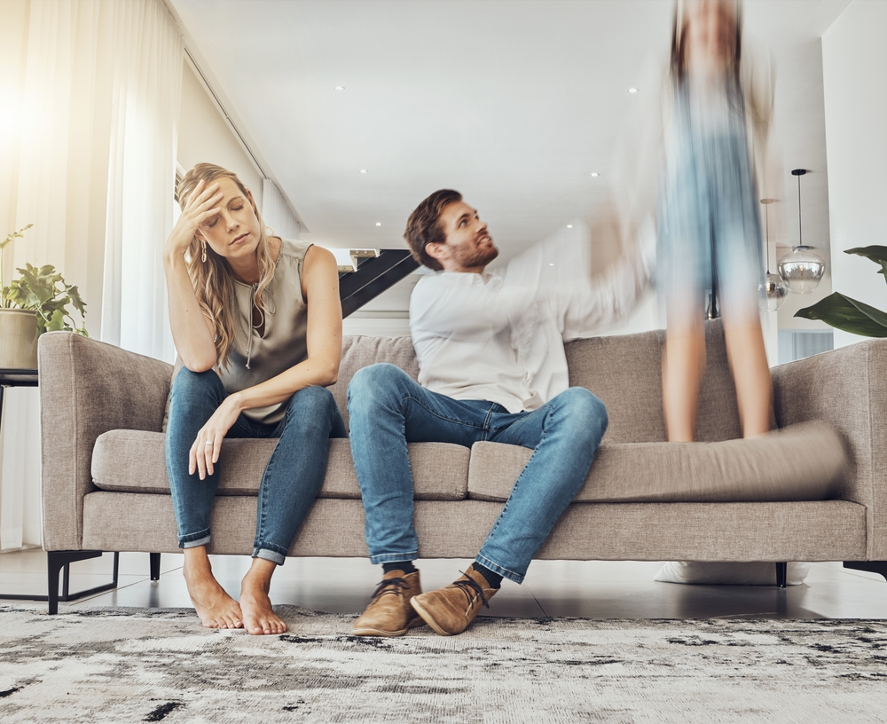 Ein Kind springt auf dem Sofa, während die nebem ihm sitzenden Eltern gestresst aussehen
