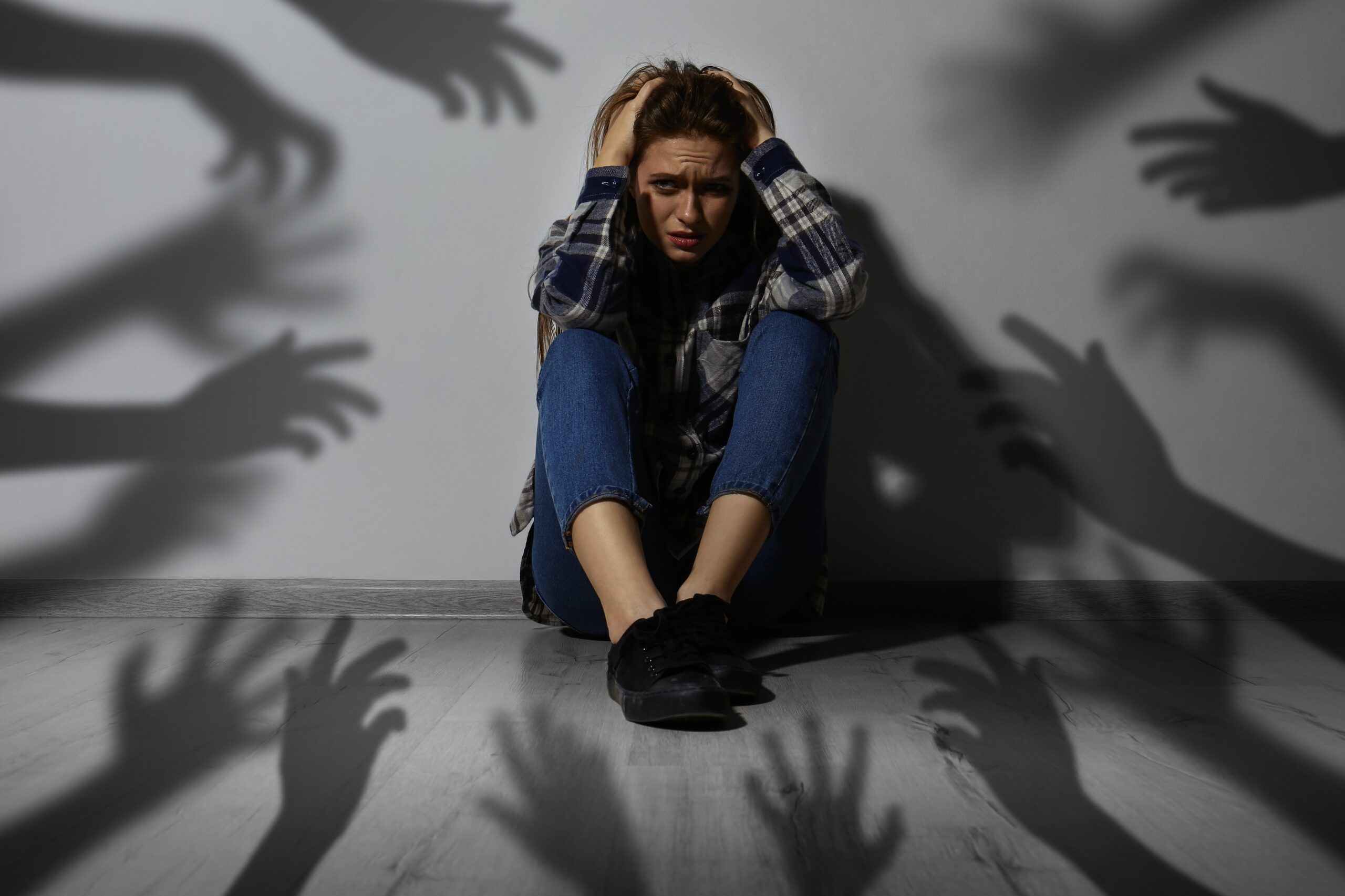 Symbolbild Pranoia - Eine Frau sitzt auf dem Boden und verzweifelt, weil Schatten von Händen nach ihr greifen.