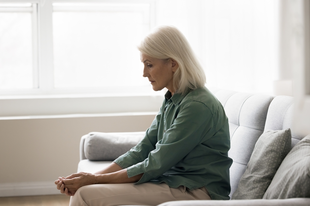 Ene ältere Person sitzt auf einem Sofa und schaut nachdenklich auf den Boden.